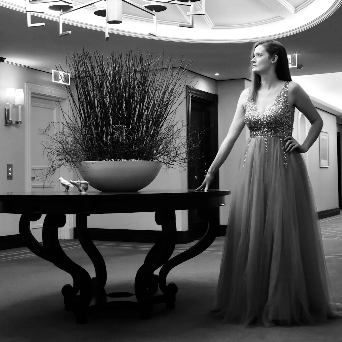 A woman in an elegant beaded evening gown in a chandelier-lit lobby.